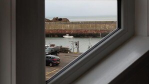 A view through a window of a coastal area with a rocky breakwater, a body of water, and parked cars. - Unique 2 Bedroom Harbour Front Apartment. License No. EL00051F.     (Dunbar)