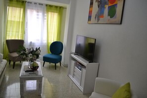 Ein Wohnzimmer mit einem weißen Sofa, einem blauen Sessel, einem Couchtisch mit Blumen und einem Flachbildfernseher auf einem Standfuß. An der Wand hängt ein Gemälde.