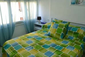 Ein Bett mit einem farbenfrohen Bettbezug mit geometrischem Muster, ein Nachttisch mit Lampe und ein Fenster mit durchsichtigen Vorhängen.