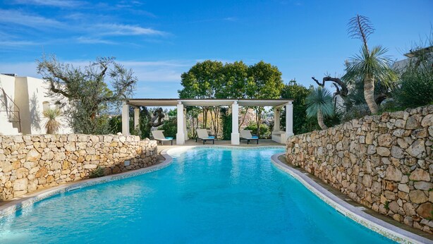 Una piscina con un muro de piedra y una zona de estar cubierta con sillas.