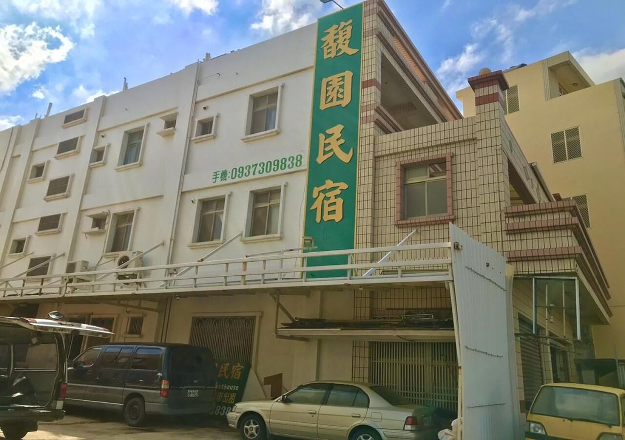 A building with a green sign indicating a guesthouse. There are multiple windows on the building, and a white car is parked in front.