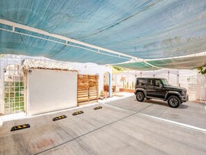 A covered parking area with a black vehicle, a building with a thatched roof, and a wooden fence.