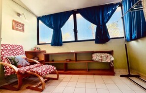 A room with a rocking chair, a bench with a floral cushion, a shelf unit, and a window with blue curtains.