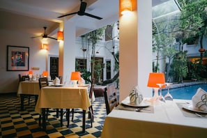 A dining area with checkered flooring, tables set with white tablecloths, orange lamps, and a view of a pool and greenery outside.