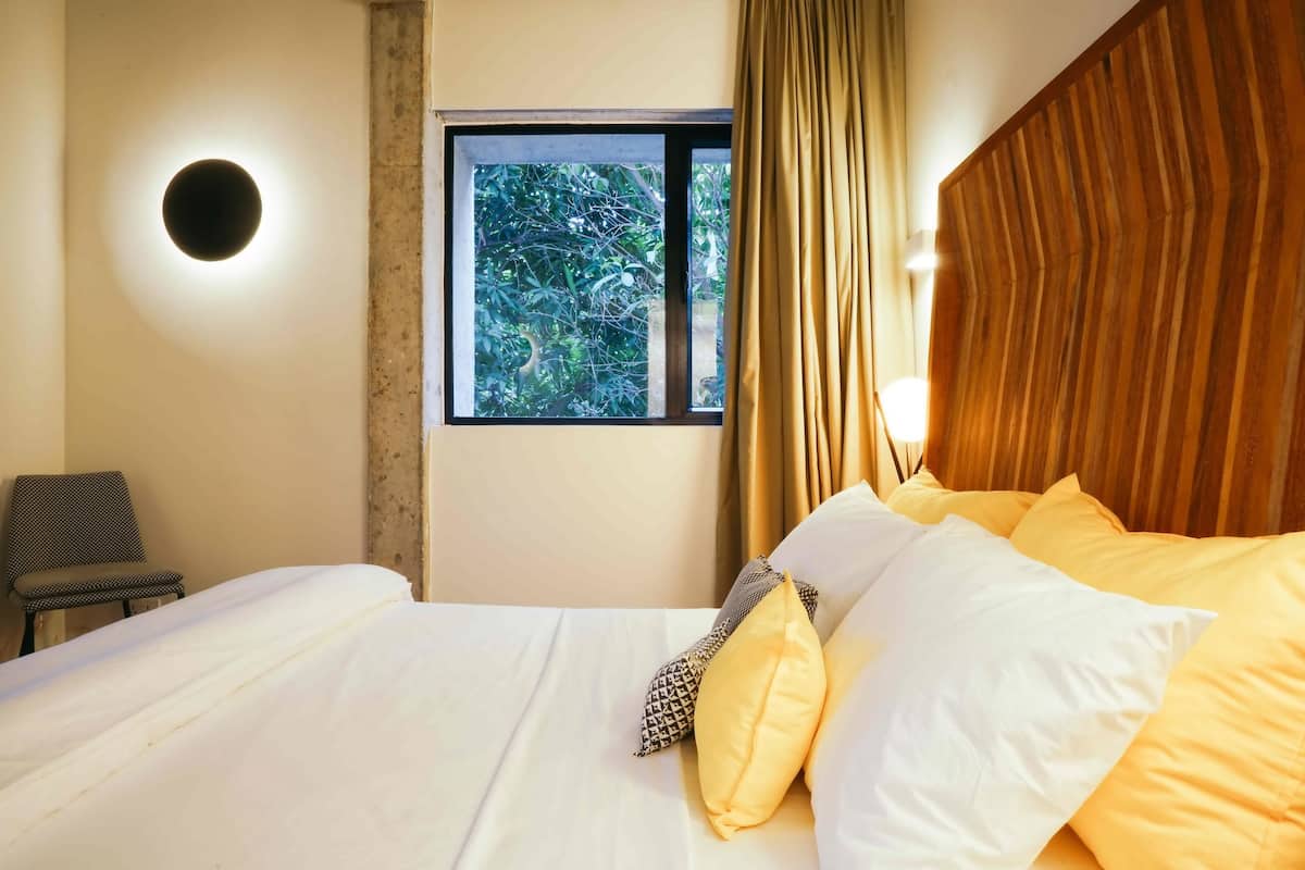 Une chambre d’hôtel avec un lit, une fenêtre donnant sur un espace verdoyant, une chaise et un luminaire fixé au mur.