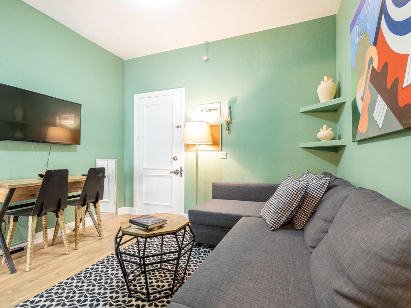 A living room with a sofa, a coffee table, and a TV. There is a door, a lamp, and a painting on the wall.