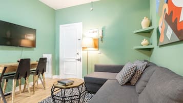A living room with a sofa, a coffee table, and a TV. There is a door, a lamp, and a painting on the wall.
