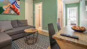 Un salon avec un canapé, une table basse et une table à manger avec des chaises. Il y a un tableau au mur, une télécommande sur la table à manger et un saladier rempli de fruits.