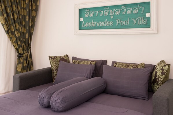 A bed with purple bedding and decorative pillows, a green sign with Thai script, and a patterned curtain.