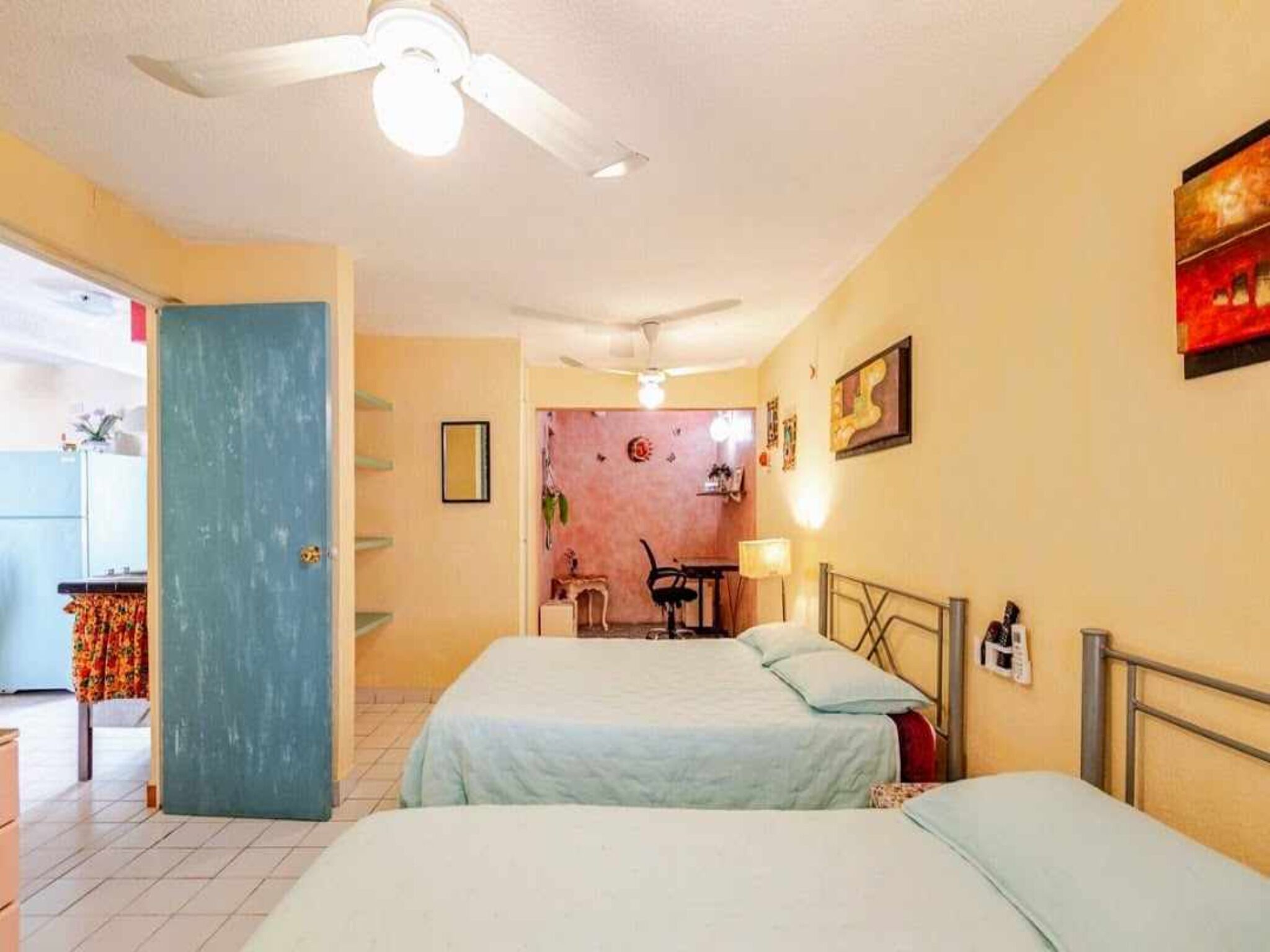 Une chambre avec deux lits, un ventilateur de plafond, une porte et, au fond, un coin cuisine.