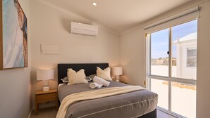 A bedroom with a large bed, two bedside tables, an air conditioner, a window with a view, and a painting on the wall.