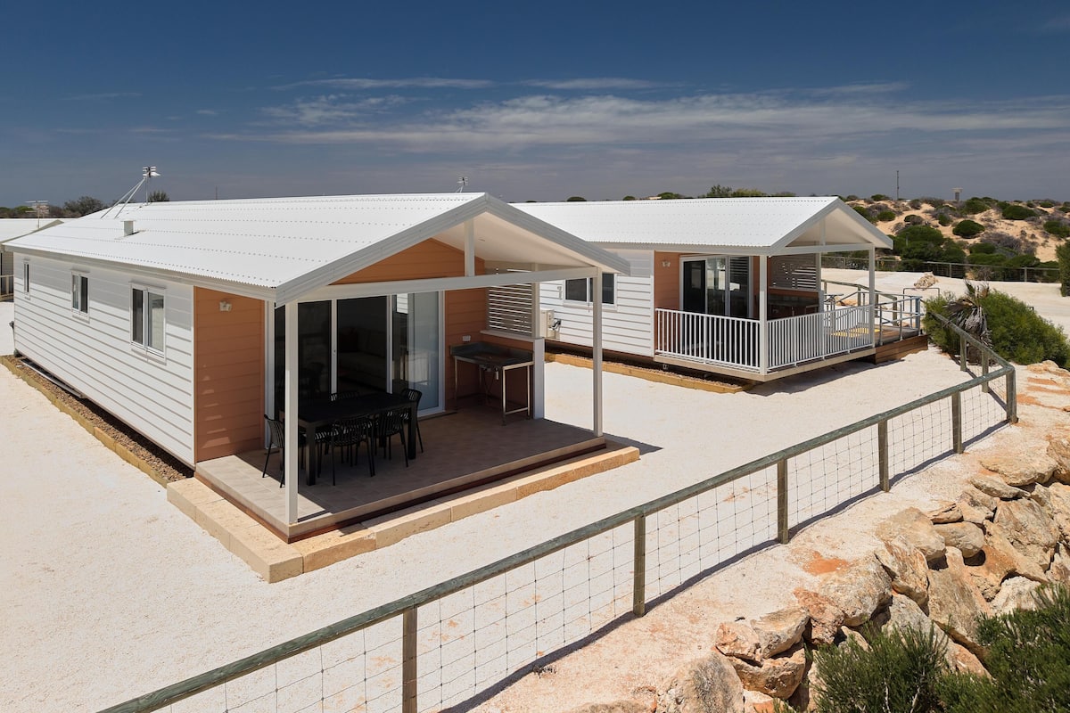 Two mobile homes with attached patios, one with a table and chairs, and the other with a balcony.
