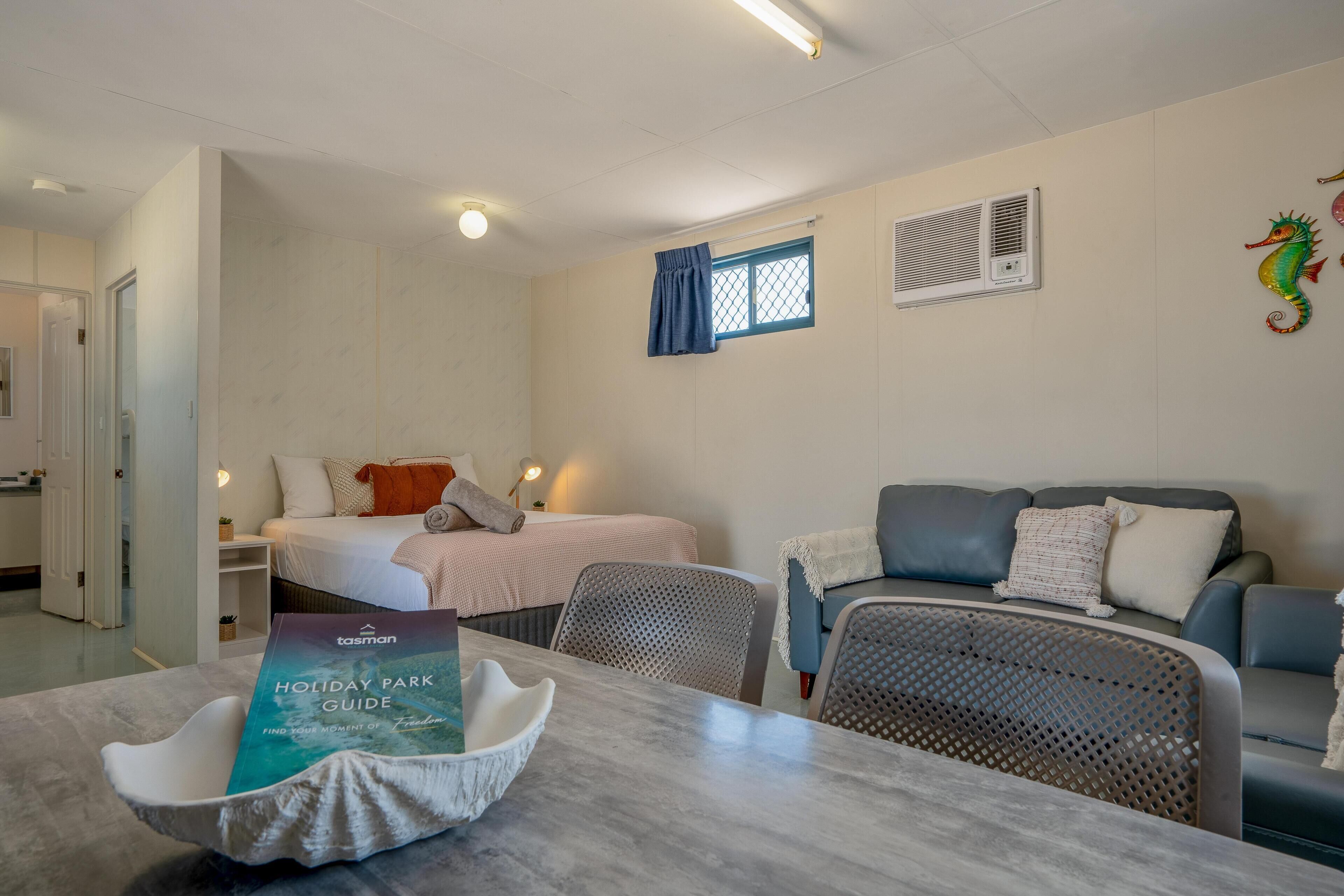 A hotel room with a bed, a sofa, a dining table, and a chair. There is a window with a curtain, a wall-mounted air conditioner, and a decorative seahorse.