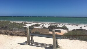 A bench with a sign that reads "BAY WATCH" overlooking a beach with a clear blue sky and calm sea.