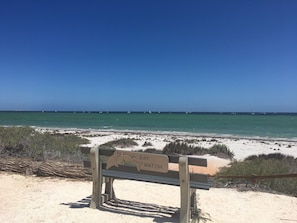 En bænk med et skilt, der står "BAY WATCH", med udsigt over en strand med en klar blå himmel og roligt hav.