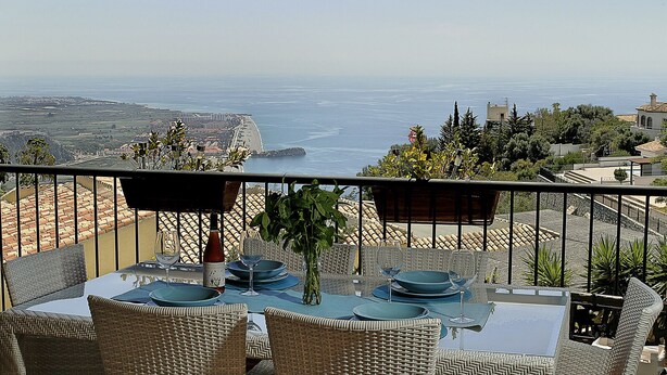 Un balcon agrémenté de meubles en osier, une table dressée pour le repas et une vue sur la mer et les bâtiments éloignés.