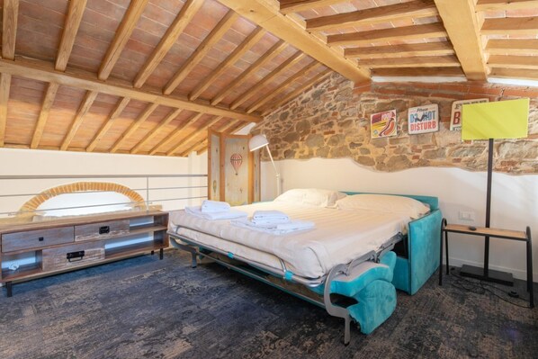 Un dormitorio con pared de piedra, techo de madera, cama con cabecera color turquesa, mesita de noche y una lámpara.
