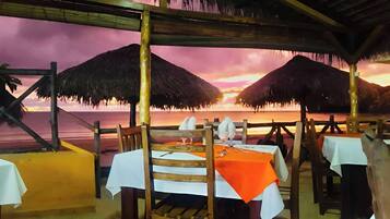 A beachside restaurant with tables and chairs under thatched umbrellas, overlooking the ocean at sunset.