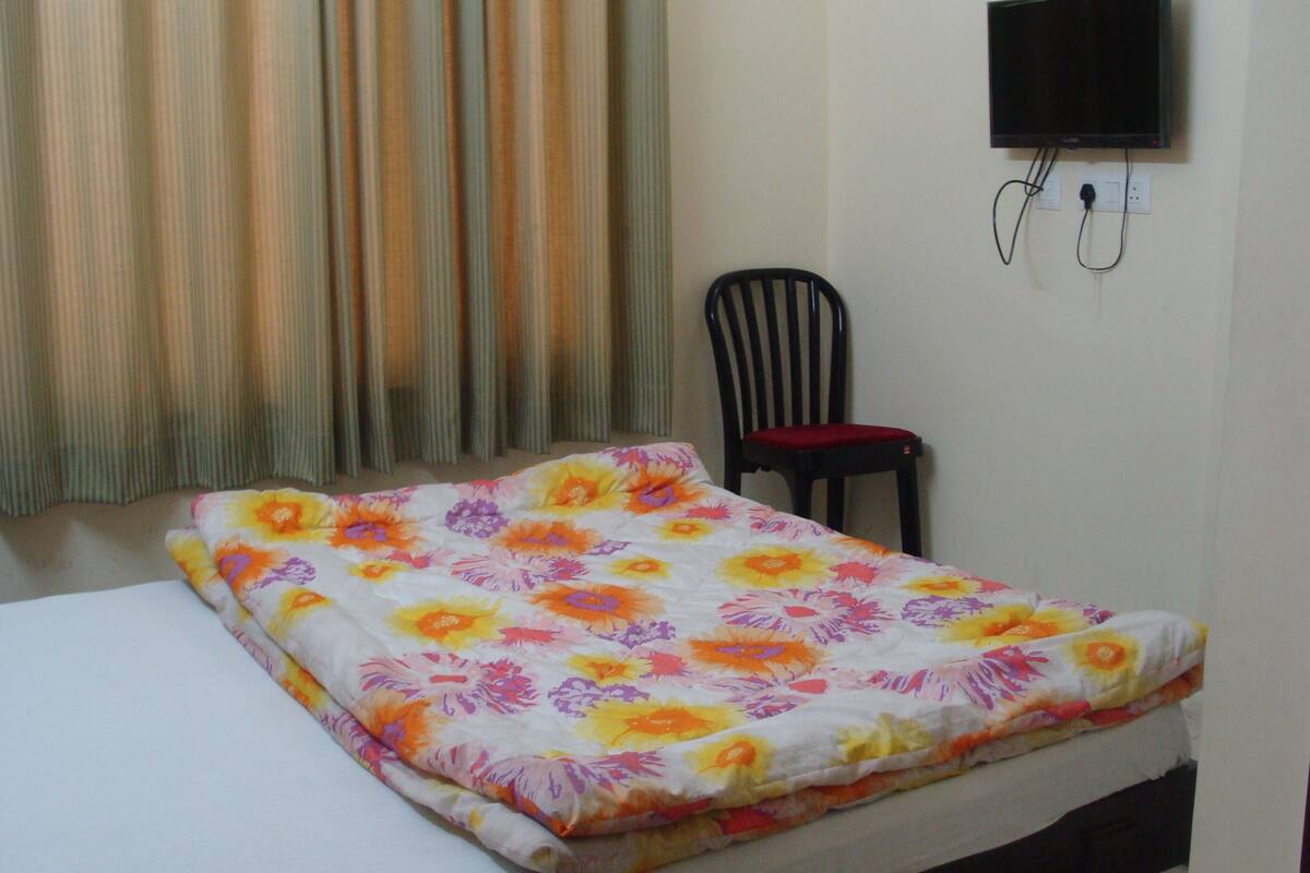 Une pièce comprenant un lit, une chaise, un téléviseur fixé au mur et une fenêtre avec des rideaux.