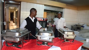 A buffet setup with stainless steel chafing dishes on a red tablecloth in a kitchen area. - Starehe link hotel (Nairobi)