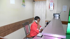 A reception desk with a person seated behind it, a clock, and a sign indicating room contacts. - Starehe link hotel (Nairobi)