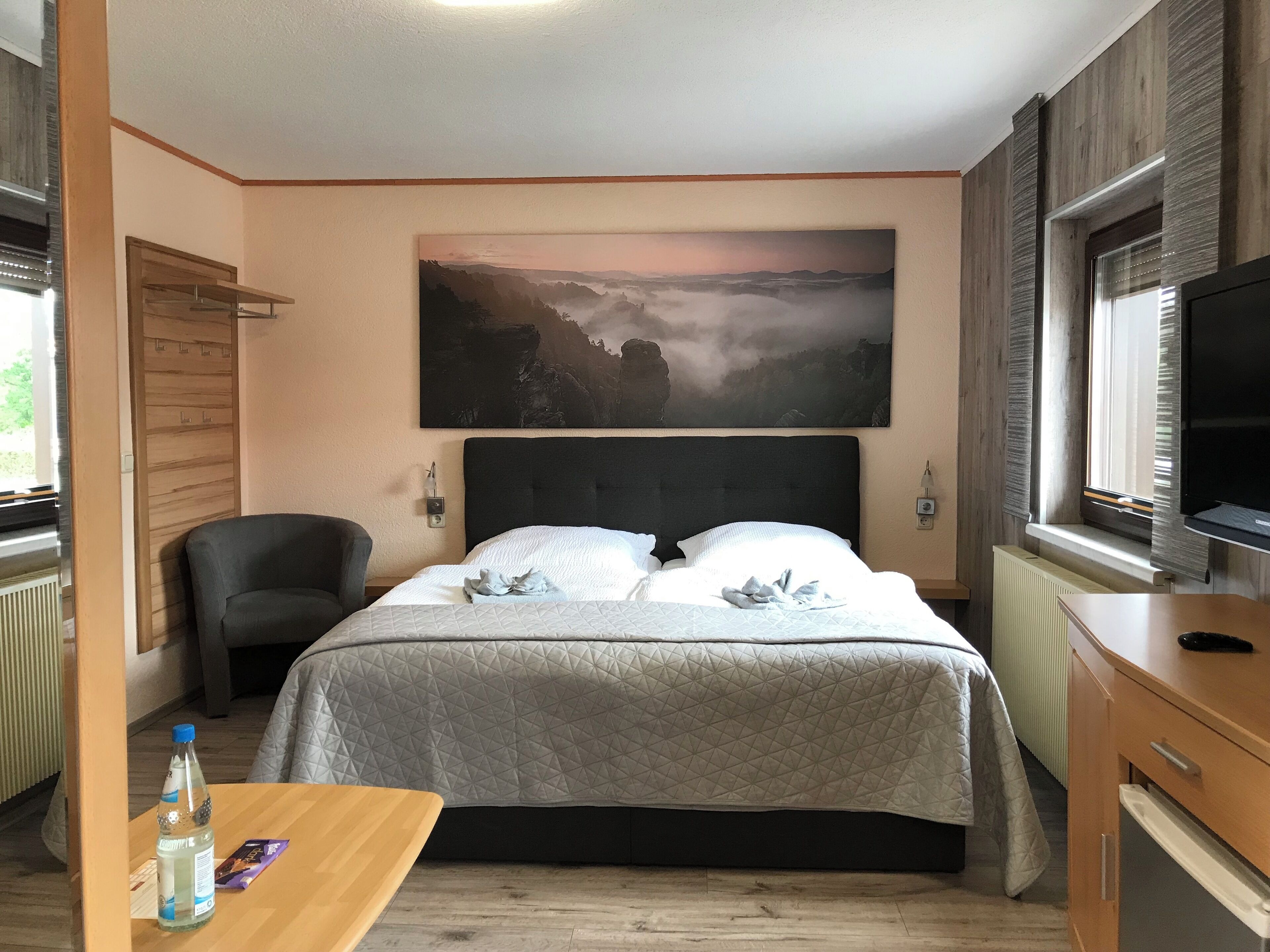 Habitación de hotel con una cama grande, una silla, una mesa con una botella y una tarjeta, un cuadro en la pared y un televisor a la derecha.