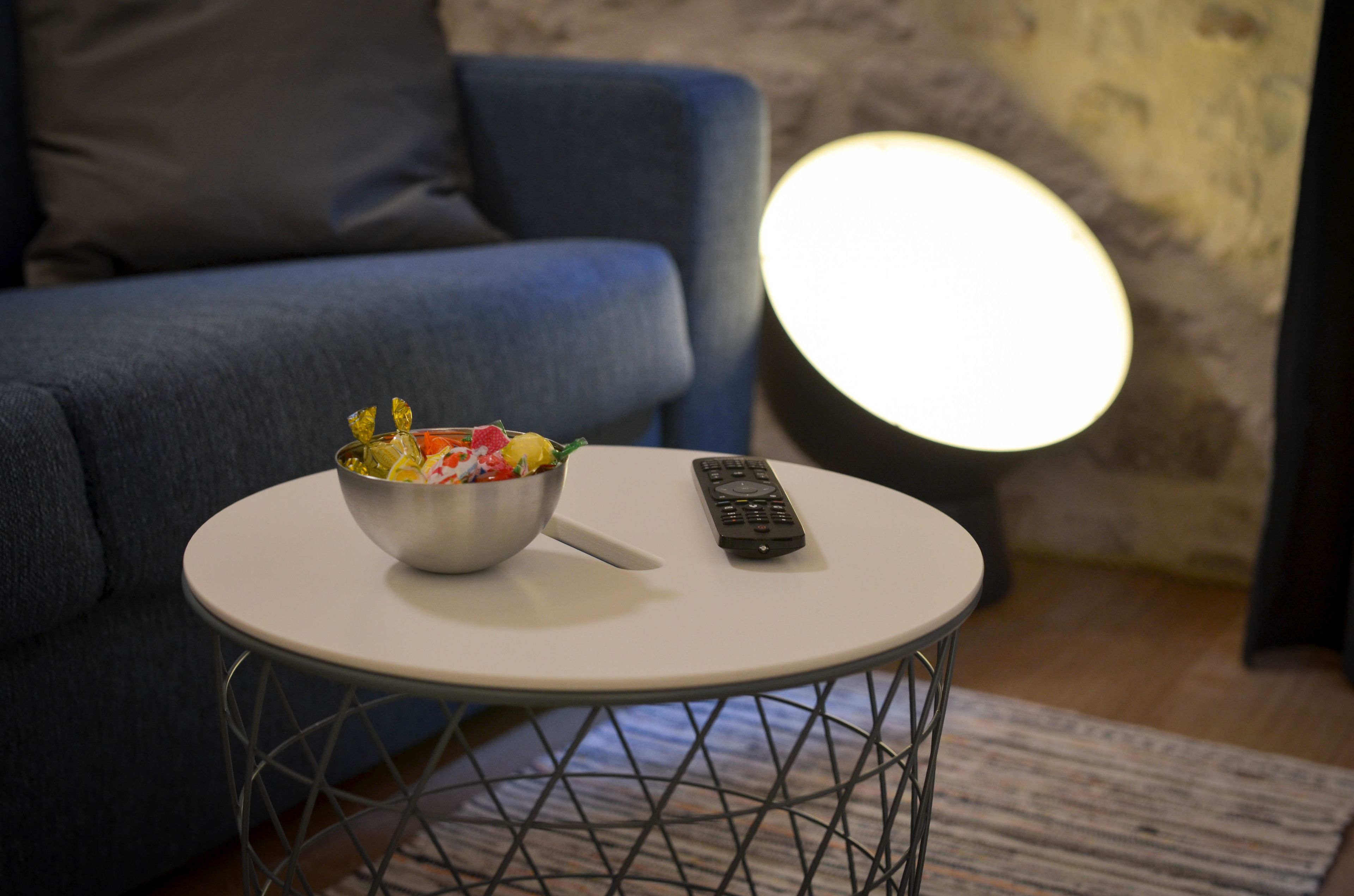 Una mesa redonda con un frutero y un control remoto, un sofá azul y una lámpara redonda en una habitación con pared texturizada.