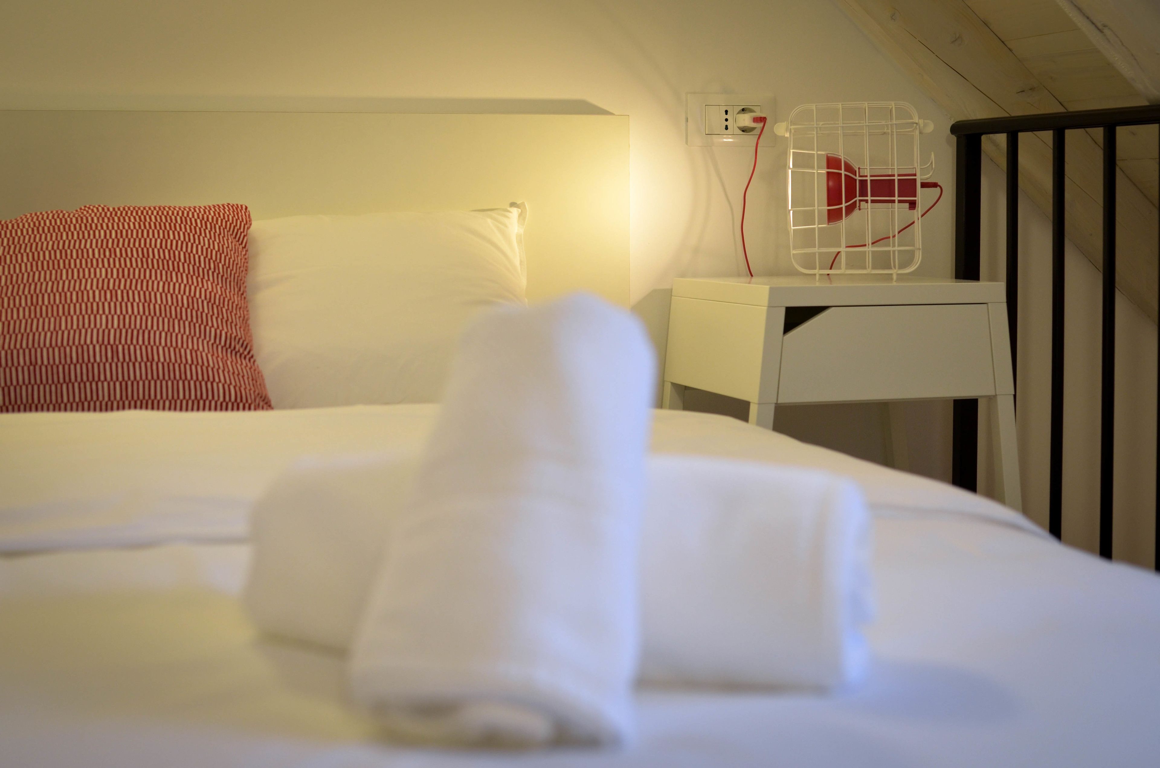 A neatly made bed with white linens and a rolled towel, a white bedside table with a red object on it, and a white wall socket.
