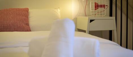 A neatly made bed with white linens and a rolled towel, a white bedside table with a red object on it, and a white wall socket.