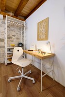 Um espaço de escritório moderno com uma secretária branca, uma cadeira de escritório branca, teto de madeira, parede de pedra e uma prateleira fixada na parede com um rádio e objetos decorativos.