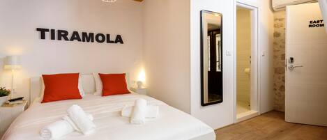 Una habitación de hotel con una cama, dos almohadas rojas, ropa de cama blanca, un espejo montado en la pared y un letrero que dice "TIRAMOLA".