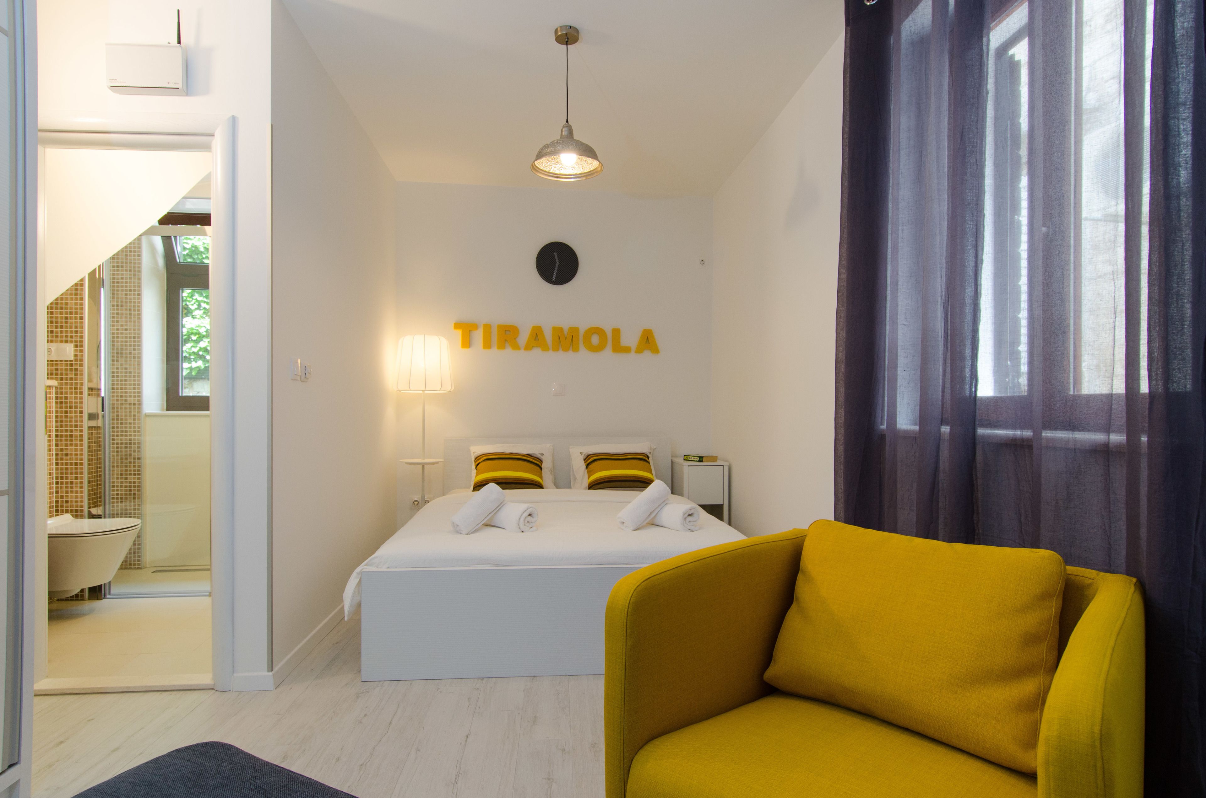 A hotel room with a bed, a yellow armchair, a bathroom with a bathtub, and a window with curtains.