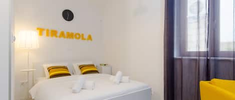 Una habitación de hotel con cama, mesita de noche, lámpara, silla y ventana con cortinas.