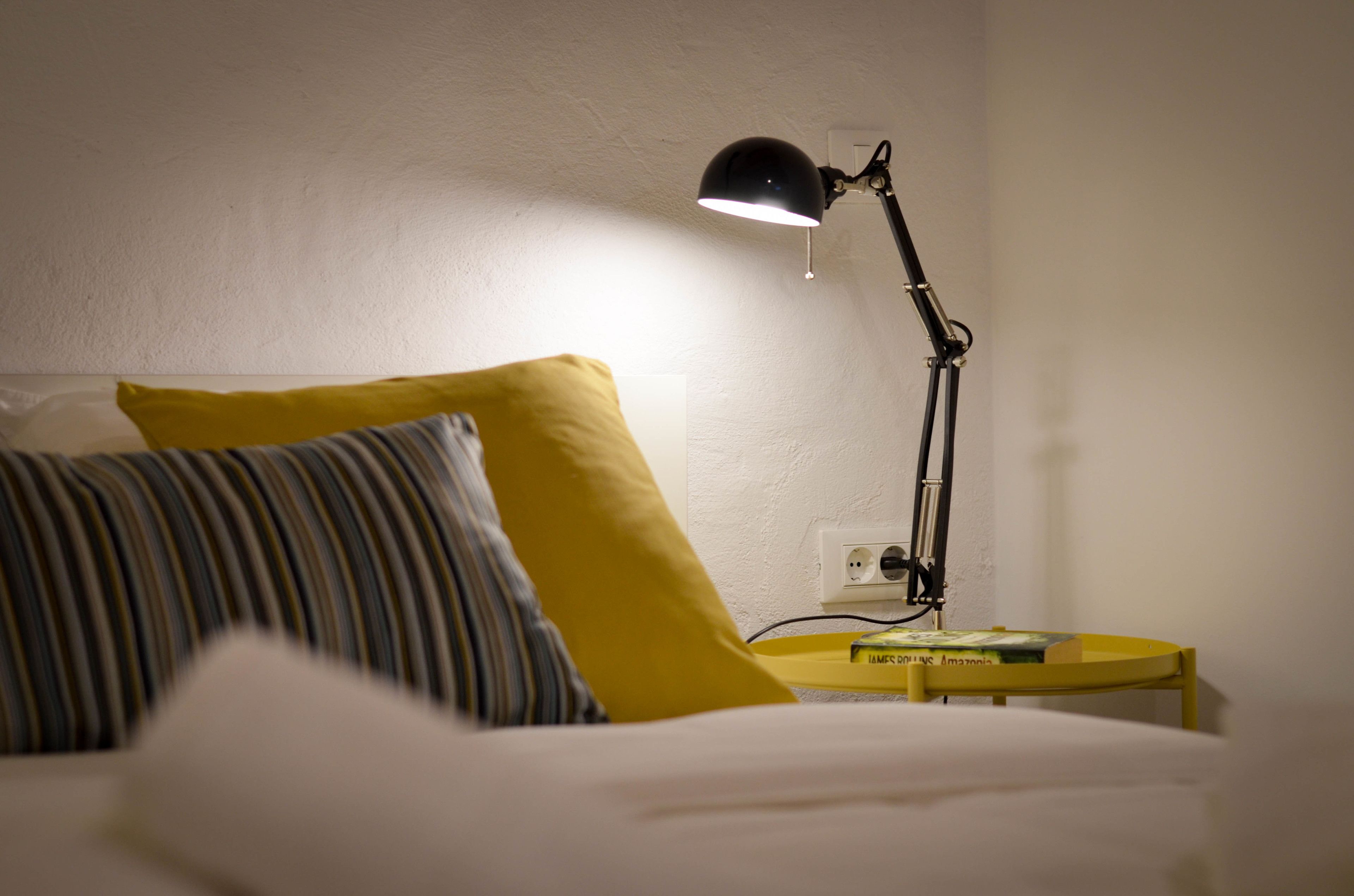 A bed with white bedding, a yellow pillow, and a black desk lamp on a yellow side table.