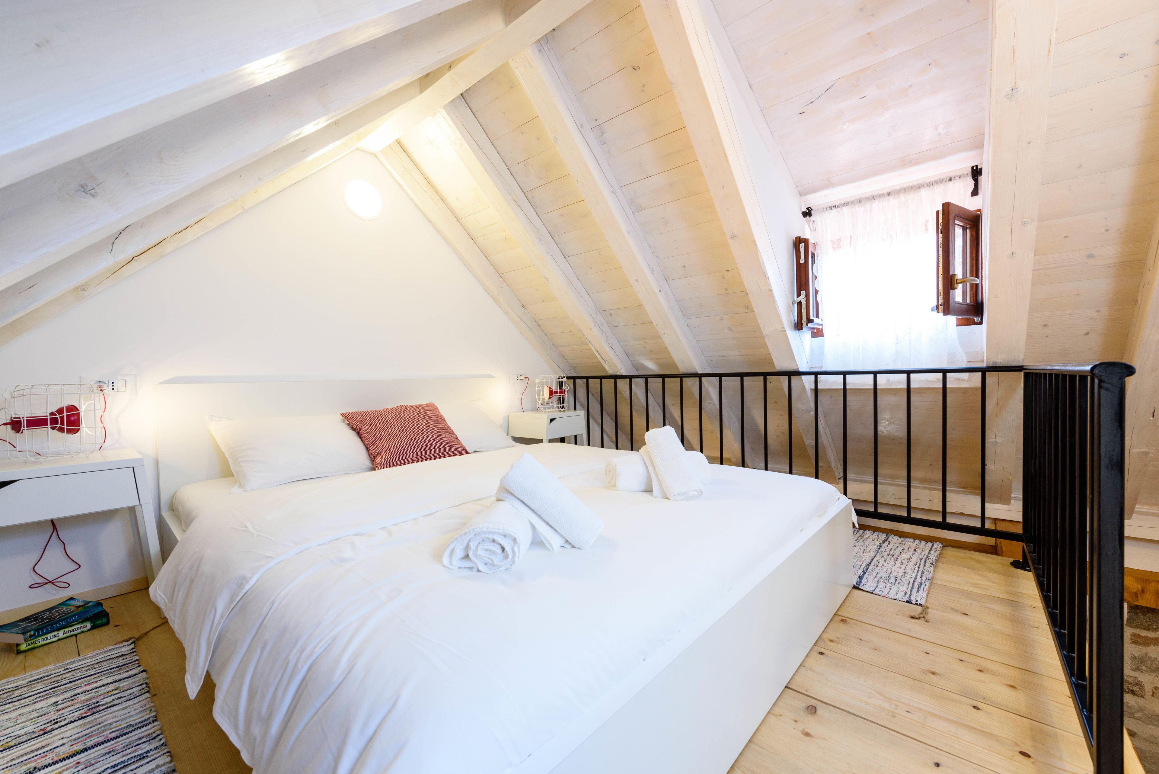 A bedroom with a sloped ceiling, a bed with white linens, a red pillow, and a nightstand with a lamp and books.