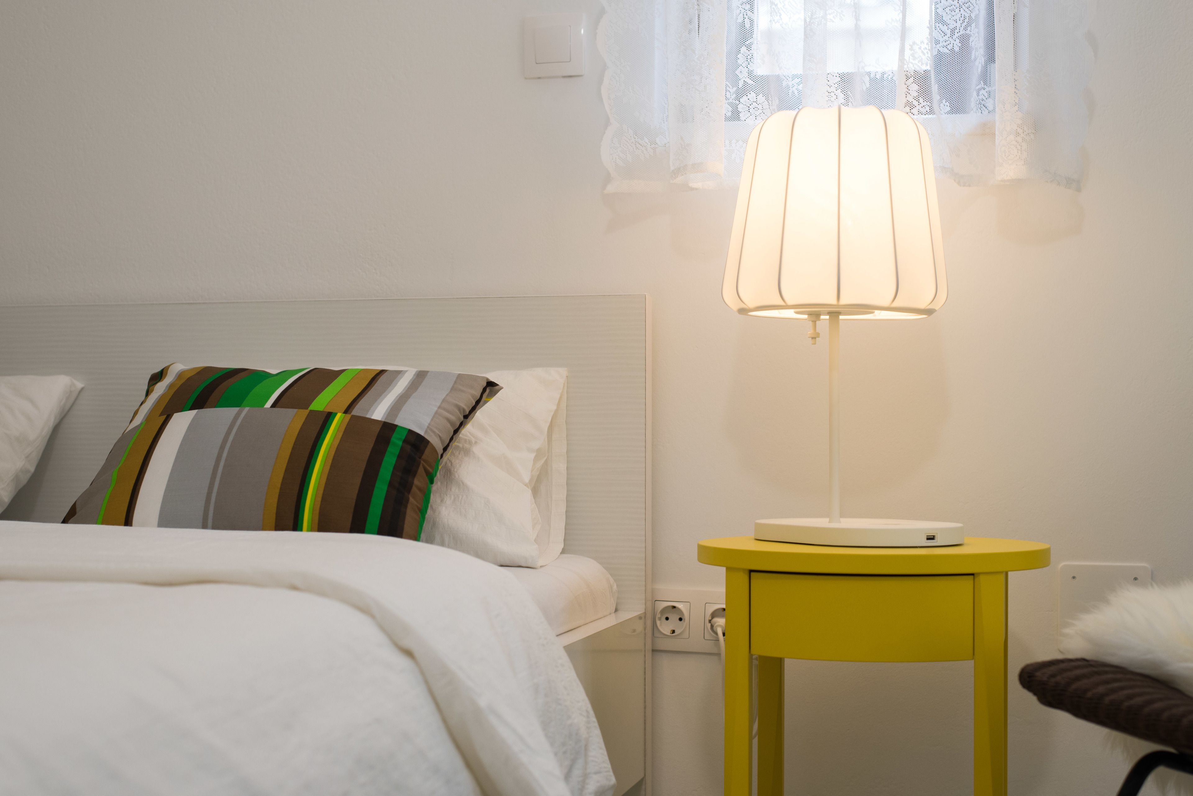 A neatly made bed with striped pillows, a bedside table with a lamp, and a power outlet on the wall.