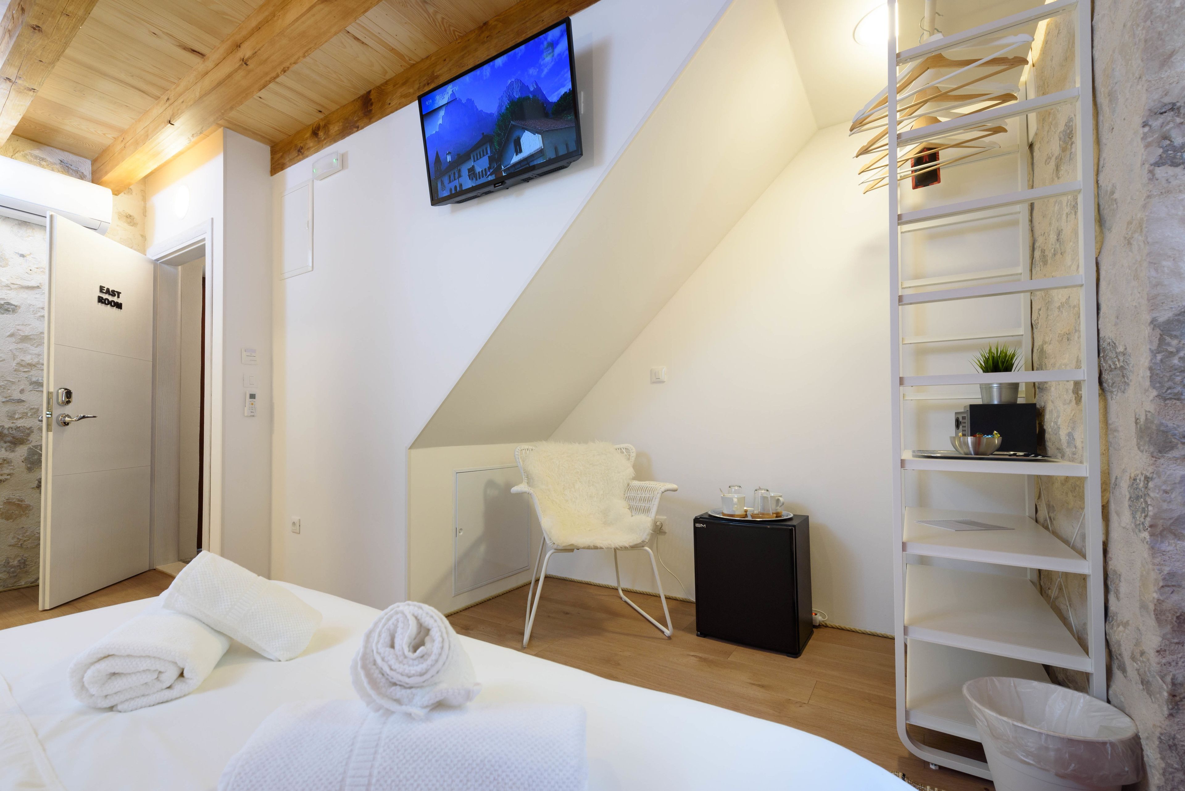 Een moderne slaapkamer met een bed, een aan de muur bevestigde televisie, een witte stoel en een houten plafond.