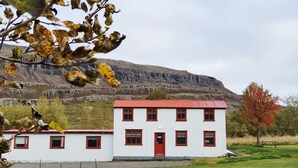 A two-story building with a red door and white walls, surrounded by a grassy field and a mountain in the background. - Fossardalur Guesthouse - Hostel (Djupivogur)