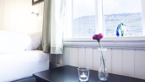 A bedroom with a bed, a nightstand, a vase with a flower, and a window with a view. - Fossardalur Guesthouse - Hostel (Djupivogur)