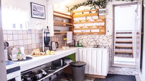 A compact kitchen with a tiled backsplash, a built-in stove, and a sink. There are shelves above the counter holding dishes and a small window with a view. - Fossardalur Guesthouse - Hostel (Djupivogur)