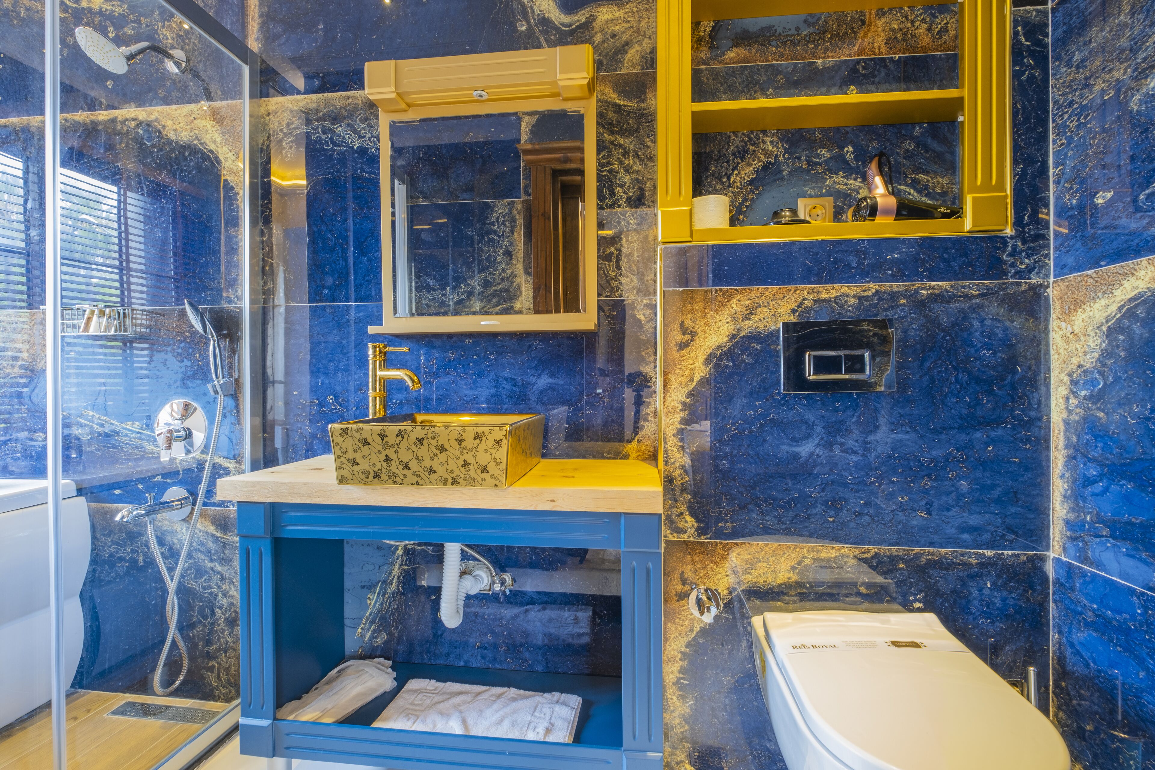 Un baño con pared de mármol azul, lavamanos blanco y zona de ducha con cerramiento de vidrio.