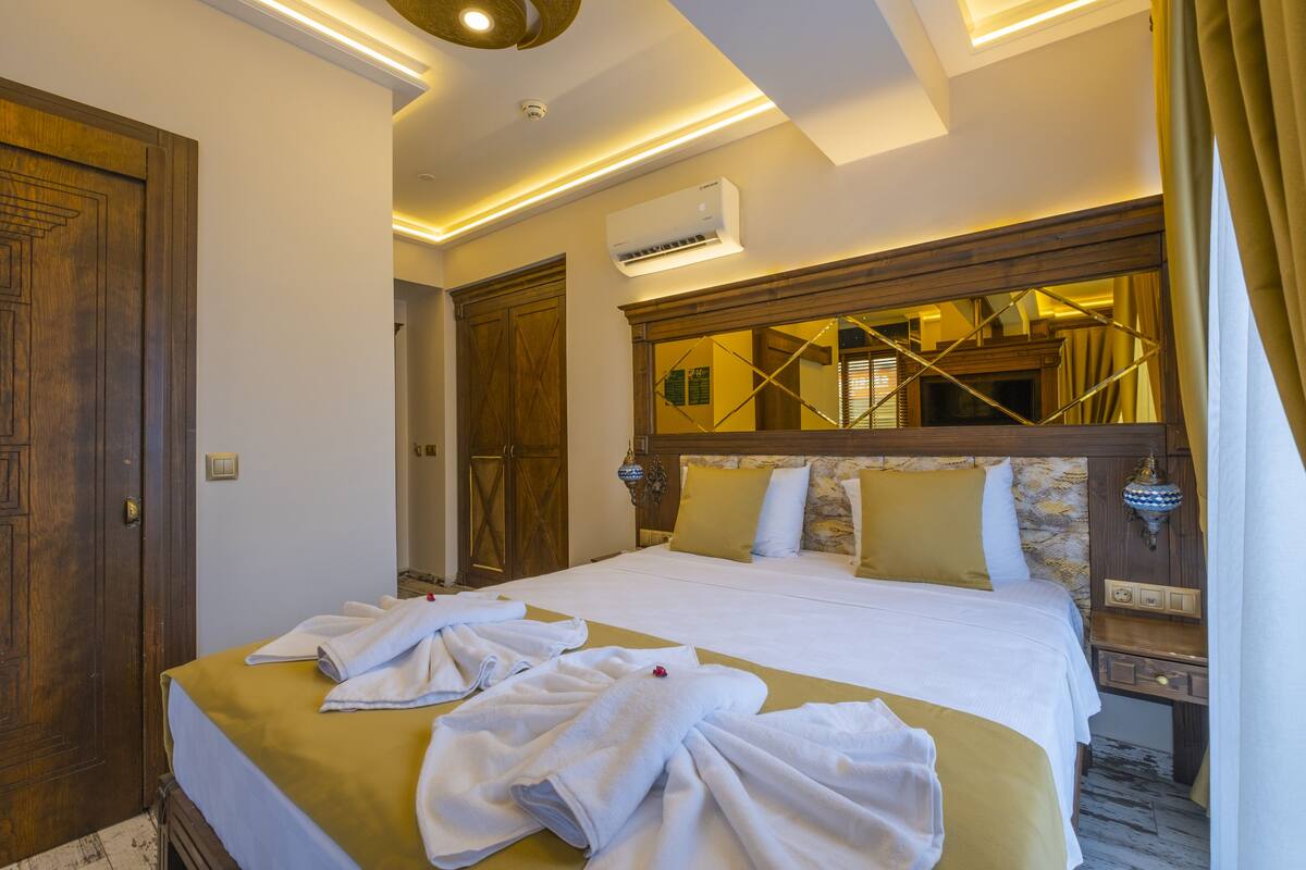 A hotel room with a large bed, a wooden wardrobe, a bedside table with a lamp, and a mirror above the bed.