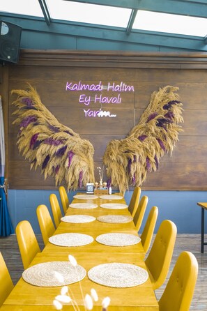 A long dining table with yellow chairs, a wooden wall with decorative feathers, and a neon sign. - Reis Royal Hotel (Fethiye)