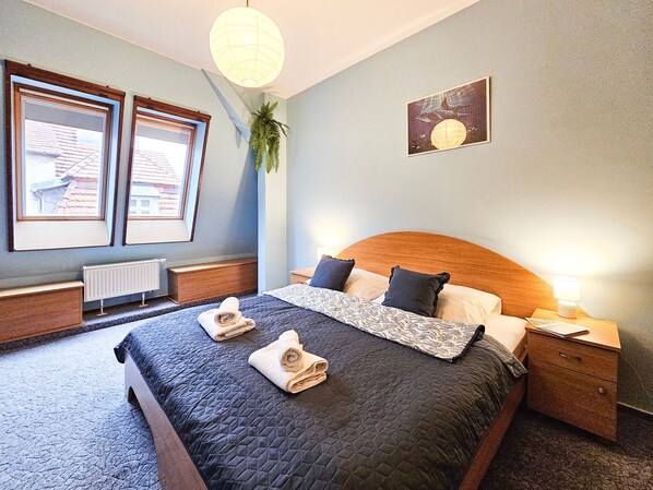 Een netjes opgemaakt bed met een gewatteerde spreidek, twee nachtkastjes, een nachtkastje met een lamp, een ingelijste foto aan de muur en een hangende plant.