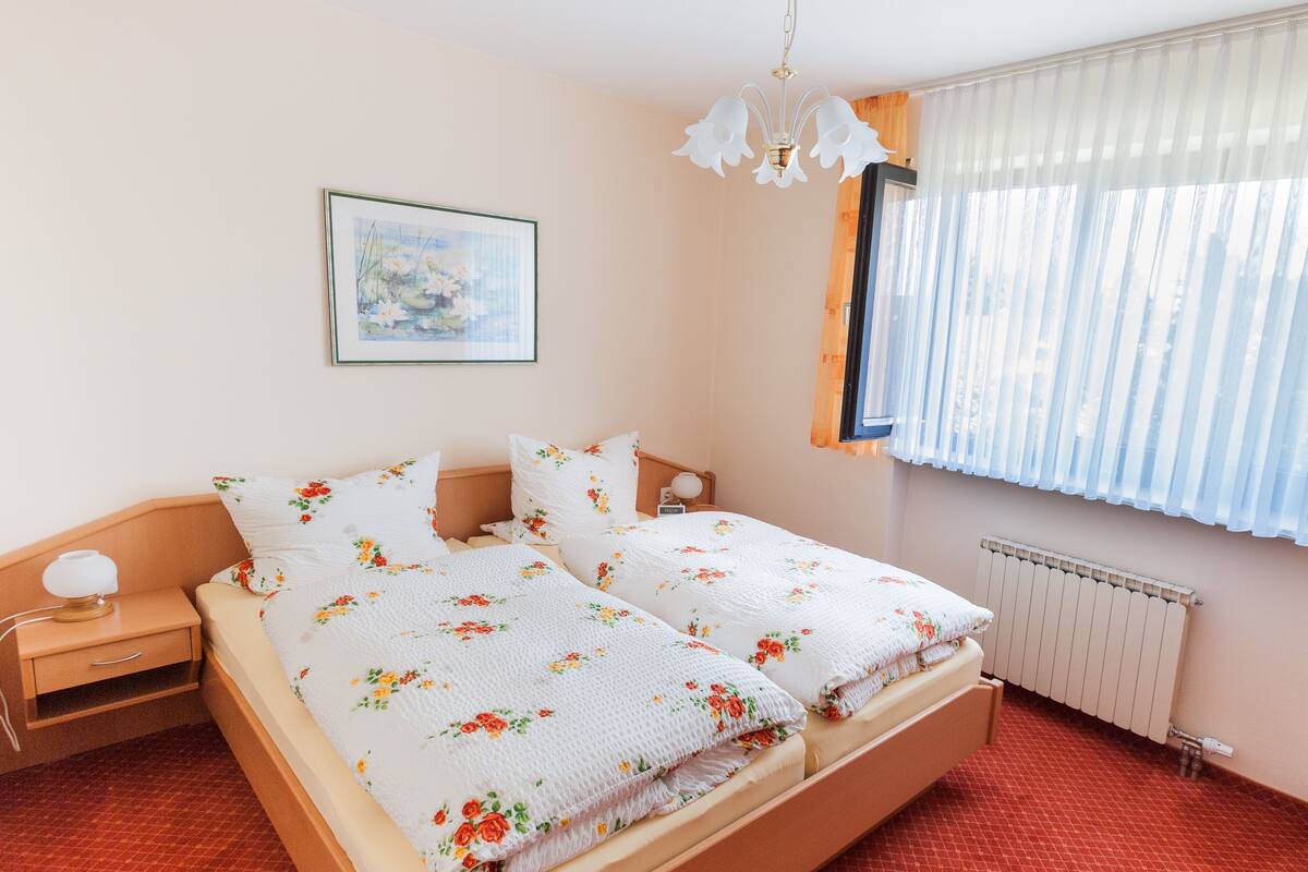 A double bed with floral bedding, a bedside table with a lamp, a framed picture on the wall, and a window with sheer curtains.