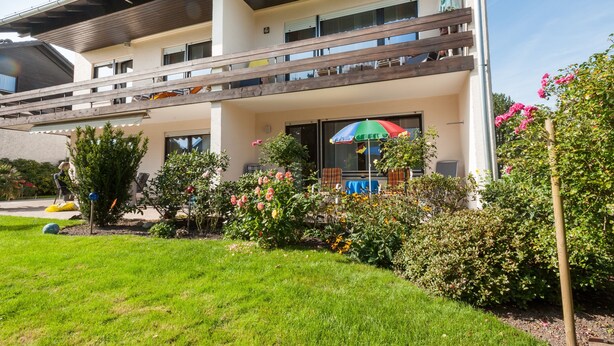 Un edificio de dos pisos con balcón, un espacio de estar al aire libre cubierto y un jardín cuidado con diversas plantas y flores.