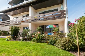 Un edificio de dos pisos con balcón, un espacio de estar al aire libre cubierto y un jardín cuidado con diversas plantas y flores.