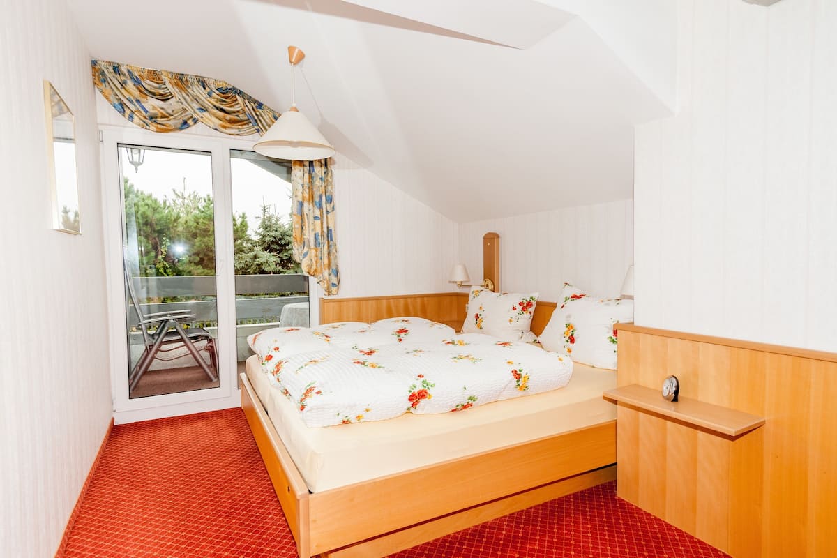 A bedroom with a wooden bed, a small bedside table, a window with curtains, and a view of greenery outside.
