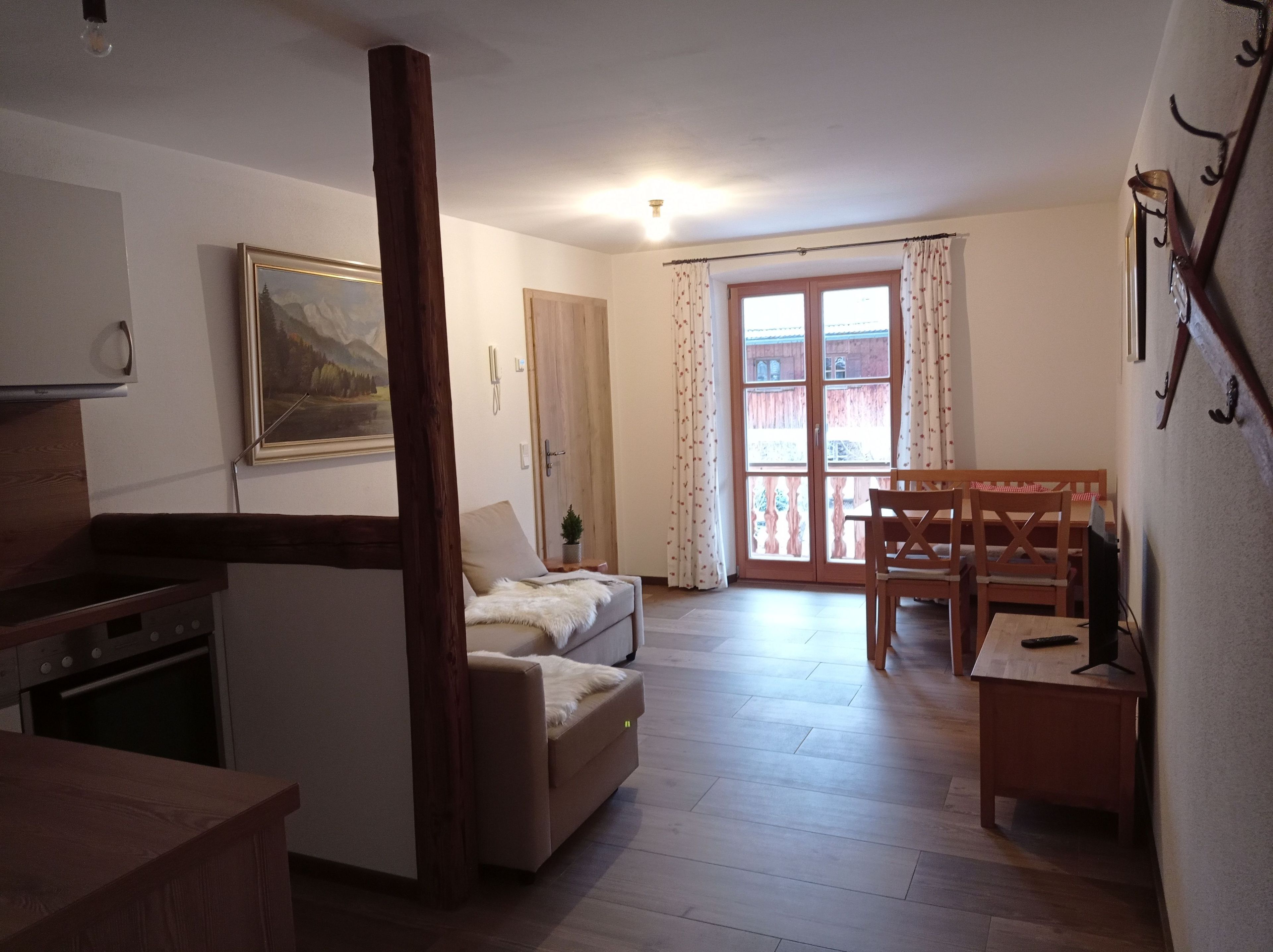 Una sala de estar acogedora con un sofá, una mesa de comedor con sillas, una viga de madera, un cuadro en la pared y una ventana con cortinas.