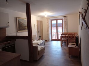A cozy living room with a sofa, a dining table with chairs, a wooden beam, a painting on the wall, and a window with curtains.